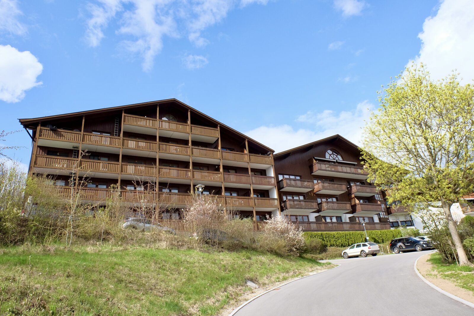 Das Apartmenthaus Himmelreich von der Zufahrt aus: vier Geschosse mit durchgehenden Holzbalkonen im alpenländischen Stil, davor die Stellplätze