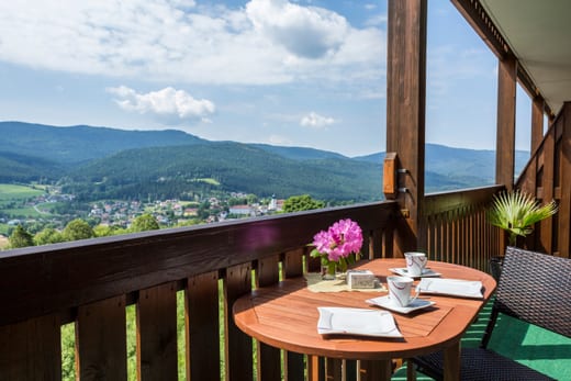 Blick vom überdachten Holzbalkon über Lam: ein gedeckter Tisch mit zwei Tassen im Vordergrund, dahinter der Ort im Tal und die bewaldeten Bergketten des Lamer Winkels