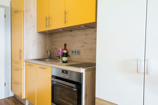 Küchenzeile mit sonnengelben Fronten, Backofen, Ceranfeld, Spüle und Hochschränken vor einer Rückwand in Holzoptik