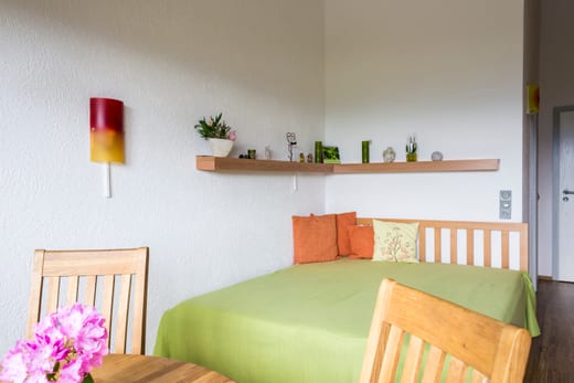 Doppelbett mit hellem Holzkopfteil, grüner Tagesdecke und orangefarbenen Kissen, darüber ein Wandregal mit Dekoration