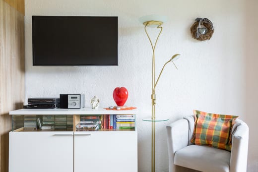 Flachbildfernseher an der Wand über einem Sideboard mit Stereoanlage, CDs und Büchern, daneben ein heller Sessel mit kariertem Kissen und eine Stehlampe