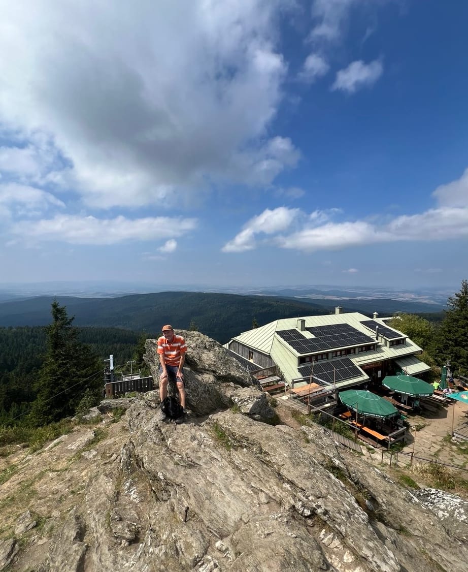 Ein Wanderer steht auf einem Felsen, dahinter eine Berghütte mit Terrasse